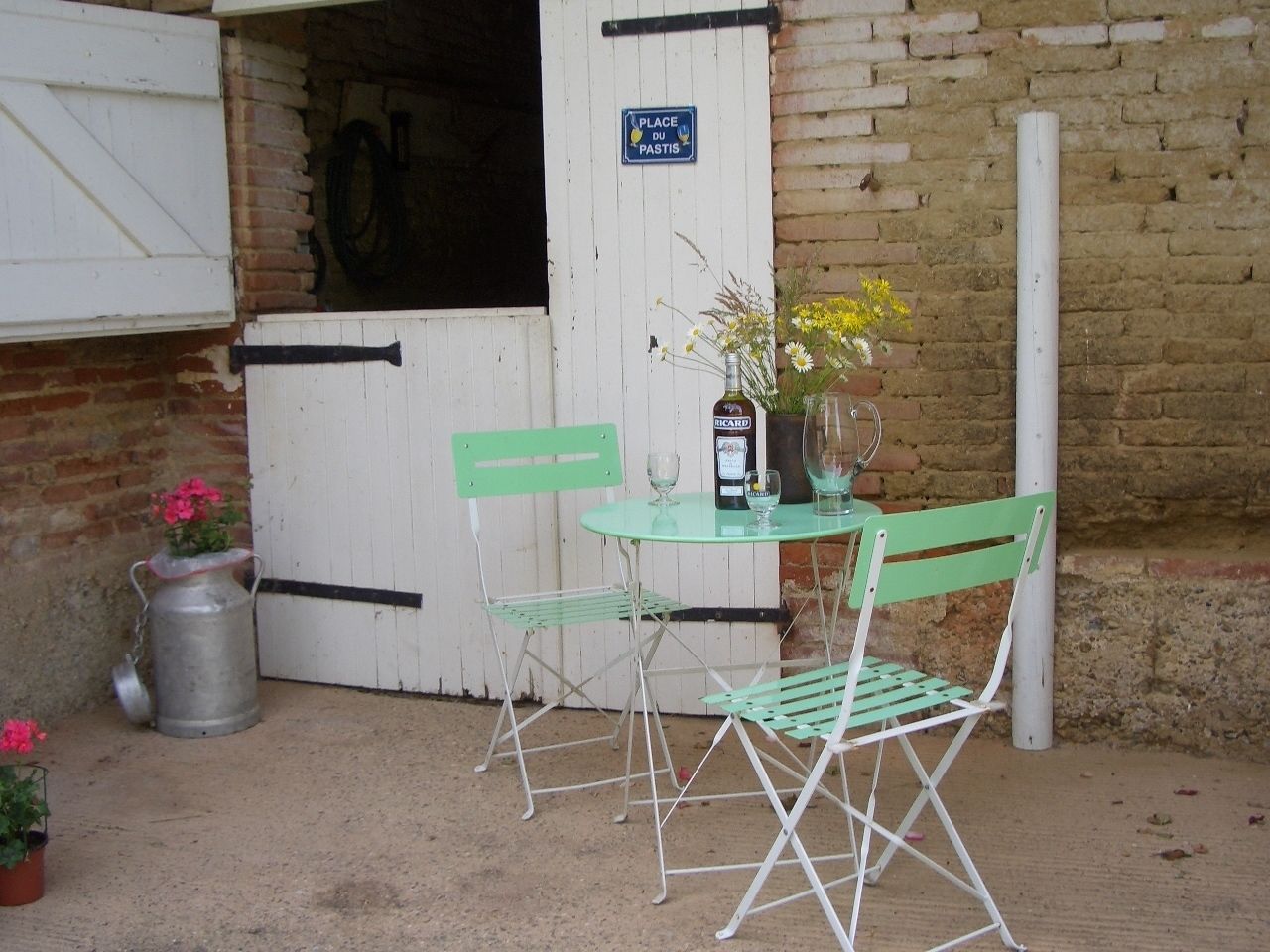 A rustic outdoor seating area with a metal table and two green chairs, a metal watering can, and a sign reading "PLACE DU PASTIS".