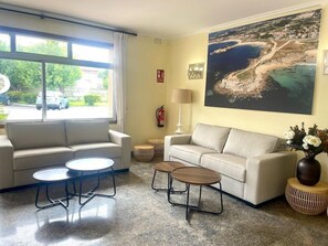 A room with two sofas, two round tables, a lamp, a fire extinguisher, and a large window with a view of a beach and buildings. - Con D´arbon (Sanxenxo)