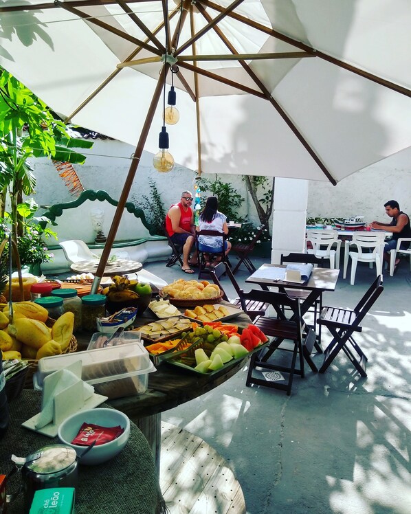 An outdoor dining area with tables and chairs, a variety of food and drinks, and a large umbrella providing shade. - Pousada Arraial Caribe (Arraial do Cabo)