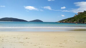 A beach with clear water, distant hills, and a building on a hill. - Pousada Arraial Caribe (Arraial do Cabo)
