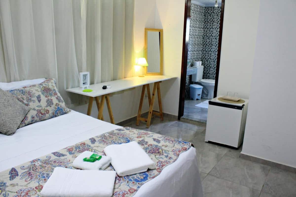 A hotel room with a bed, a desk, a mirror, a lamp, a trash bin, and a bathroom visible through an open door.