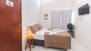 A bedroom with a bed, a bedside table with a lamp, a TV mounted on the wall, and a fan on a side table. - Pousada Arraial Caribe (Arraial do Cabo)