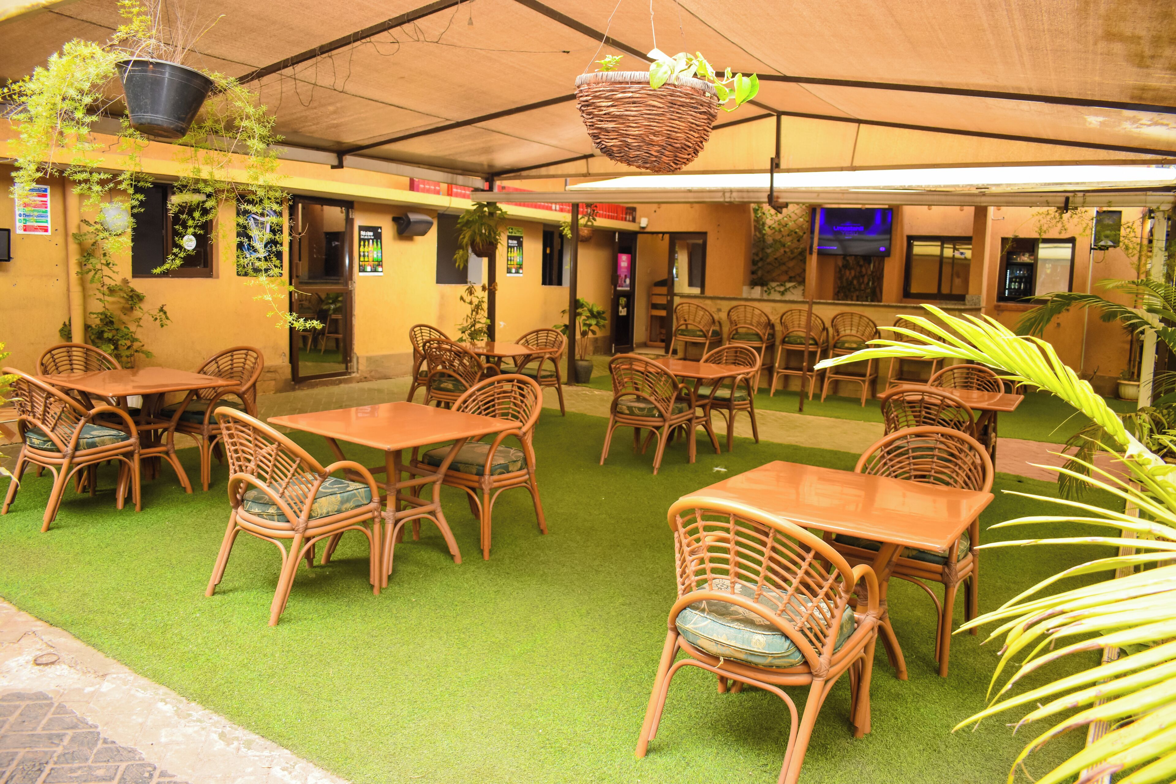 Un espace extérieur aménagé avec des tables et des chaises en osier, un revêtement de sol en gazon synthétique, des plantes suspendues et un écran de télévision fixé au mur.