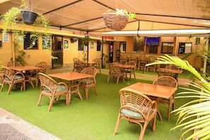 An outdoor seating area with wicker tables and chairs, artificial grass flooring, hanging plants, and a television screen on the wall. - Joy Palace (Nairobi)