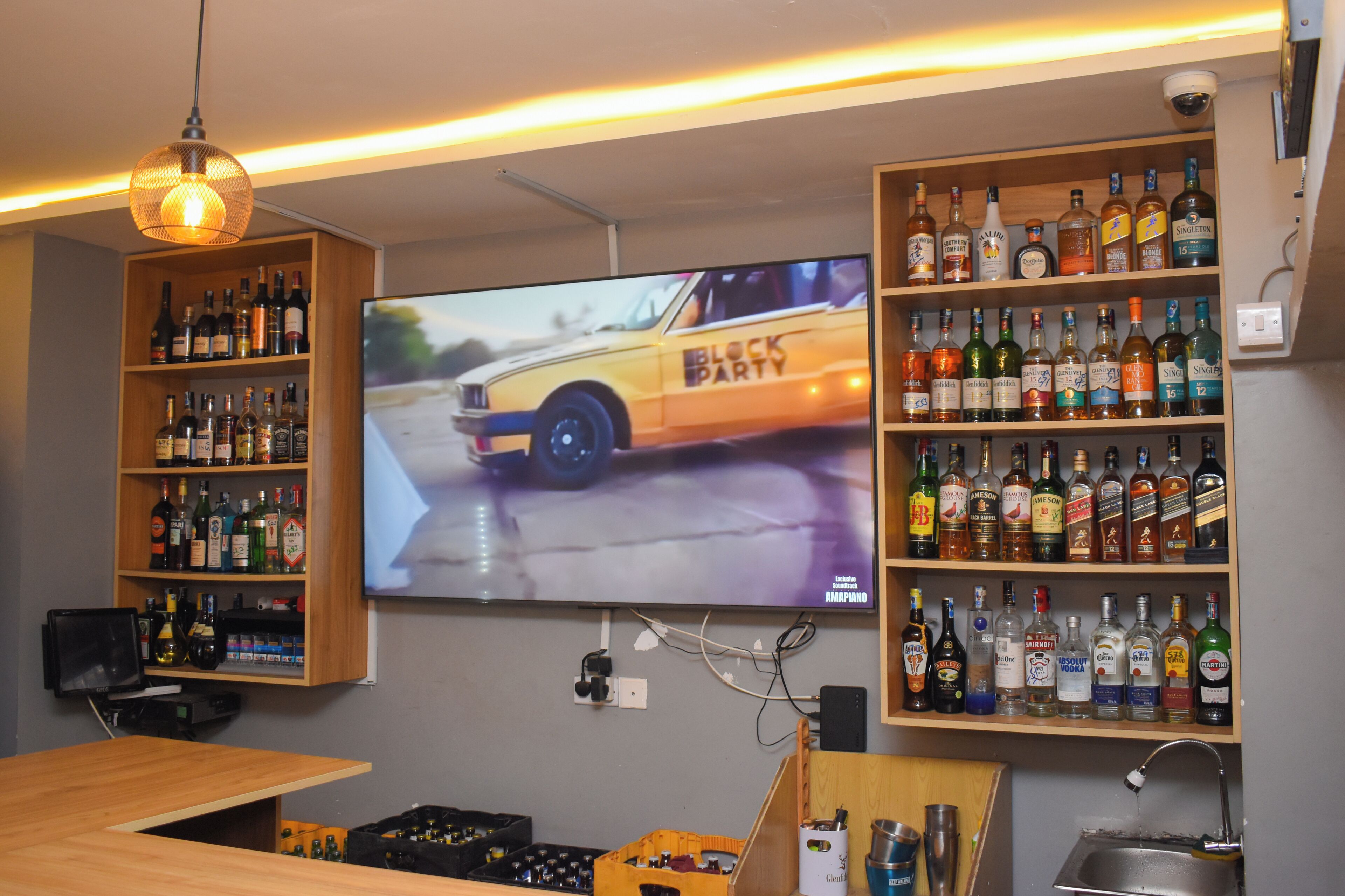 Un coin bar avec des étagères remplies de bouteilles d’alcool, un téléviseur diffusant des images de voitures et un luminaire suspendu.