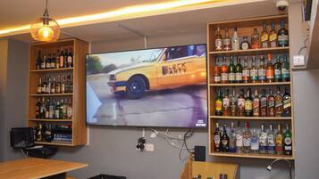 Un coin bar avec des étagères remplies de bouteilles d’alcool, un téléviseur diffusant des images de voitures et un luminaire suspendu.