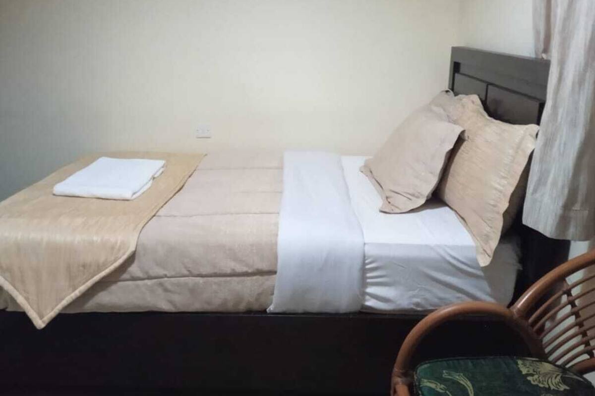A single bed with a beige bedspread and white sheets, a wooden headboard, and a green cushion on a wicker chair.