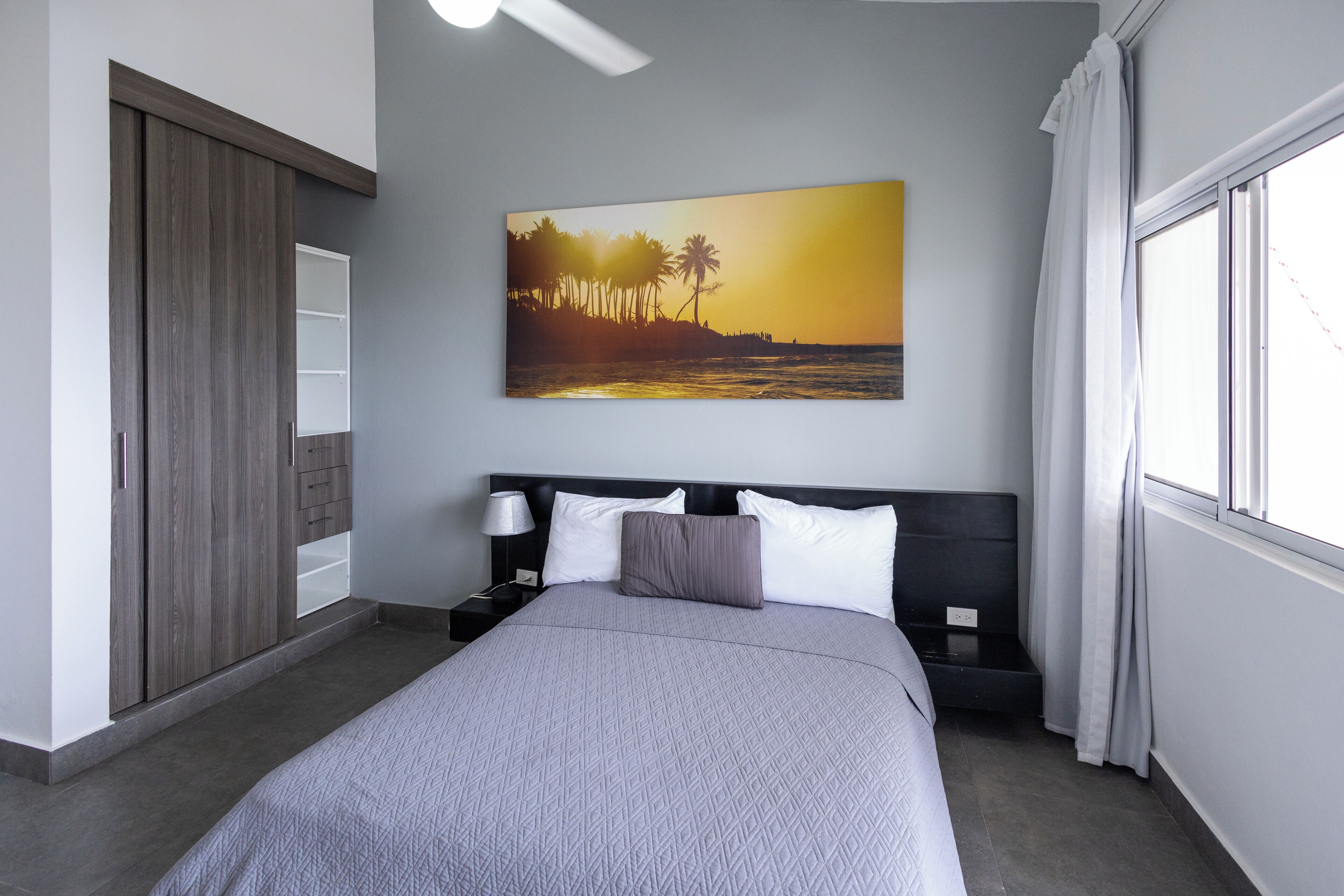 A bedroom with a bed, nightstands, a wall-mounted painting, a window with curtains, and a ceiling fan.