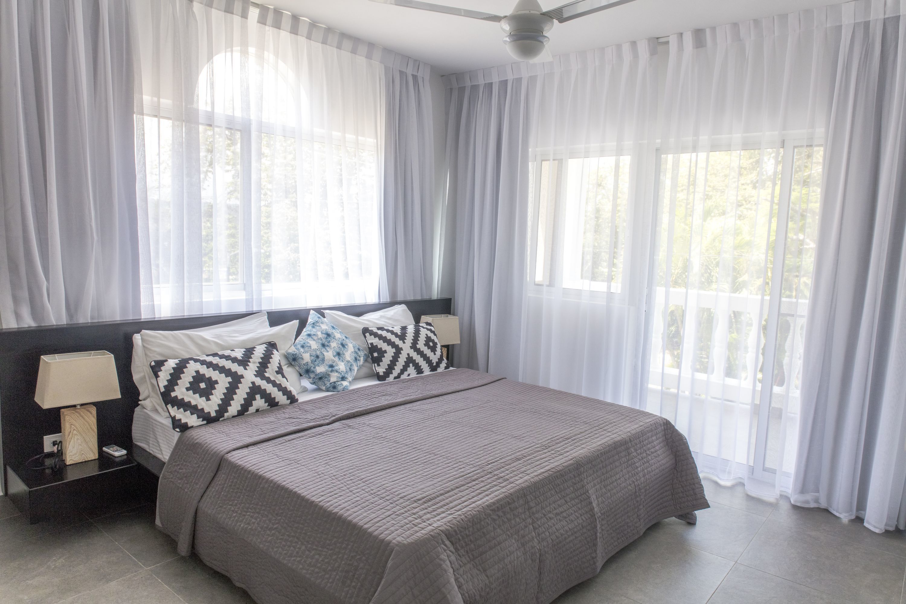 Una cama bien hecha con cubrecama y almohadas grises, una mesita de noche con lámpara y cortinas translúcidas en ventanas amplias.