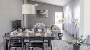 A modern dining area with a wooden table set for four, a ceiling fan, and a large window with curtains. - Ocean Palms Residences (Cabarete)
