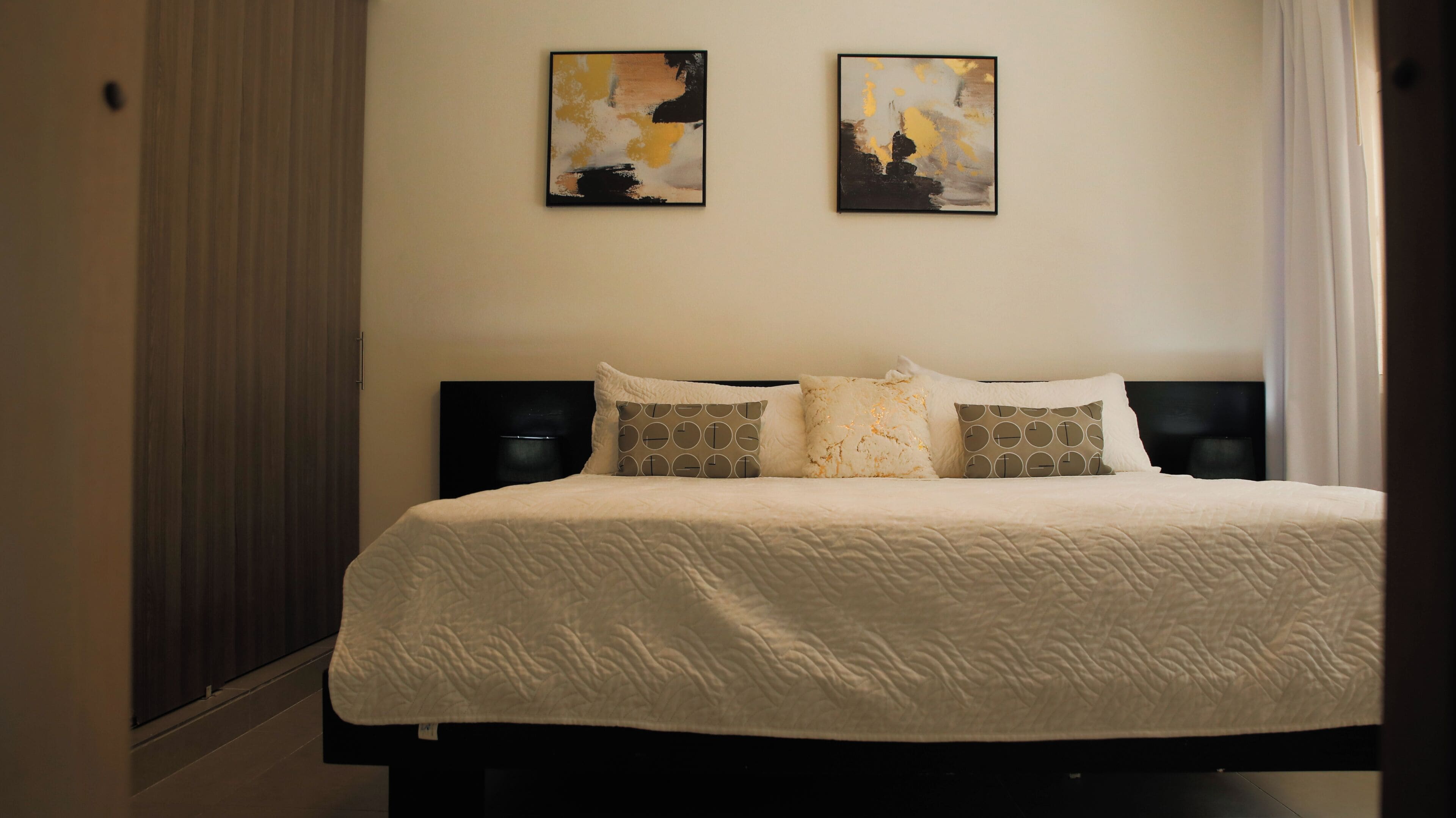 Una cama bien hecha con edredón blanco, dos almohadas con estampado geométrico y dos cuadros abstractos en la pared.