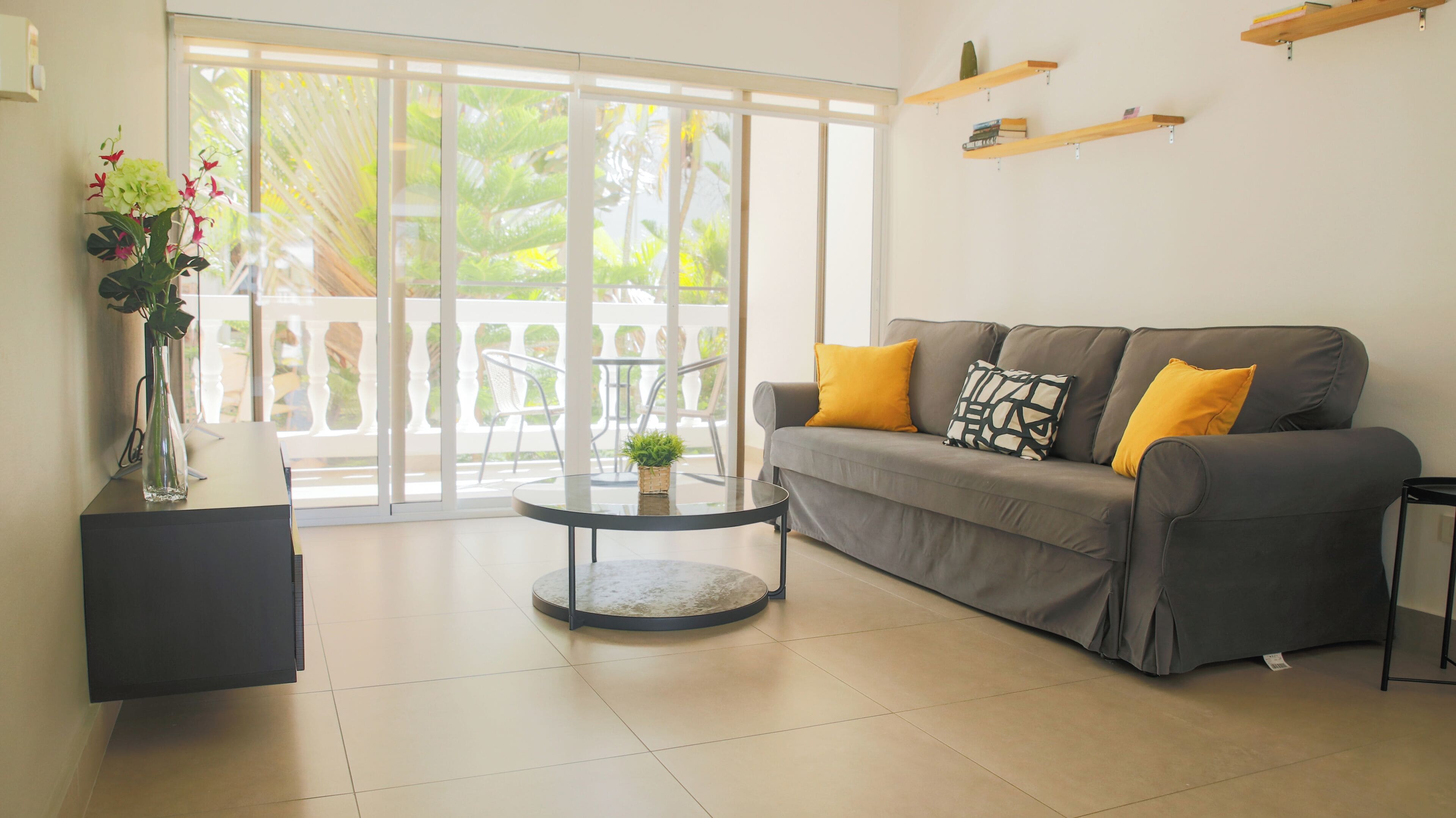 Una sala de estar moderna con un sofá gris, cojines amarillos, una mesa de centro de vidrio y una planta en maceta.