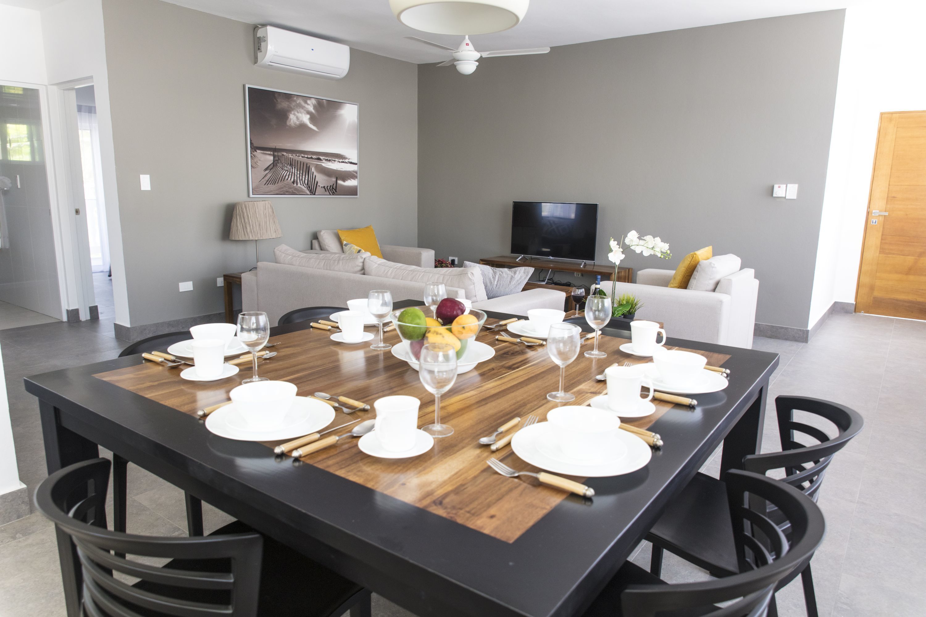 Een moderne eetruimte met een houten tafel voor zes personen, omringd door zwarte stoelen. Aan de muur tegenover de eethoek is een flatscreen televisie bevestigd.