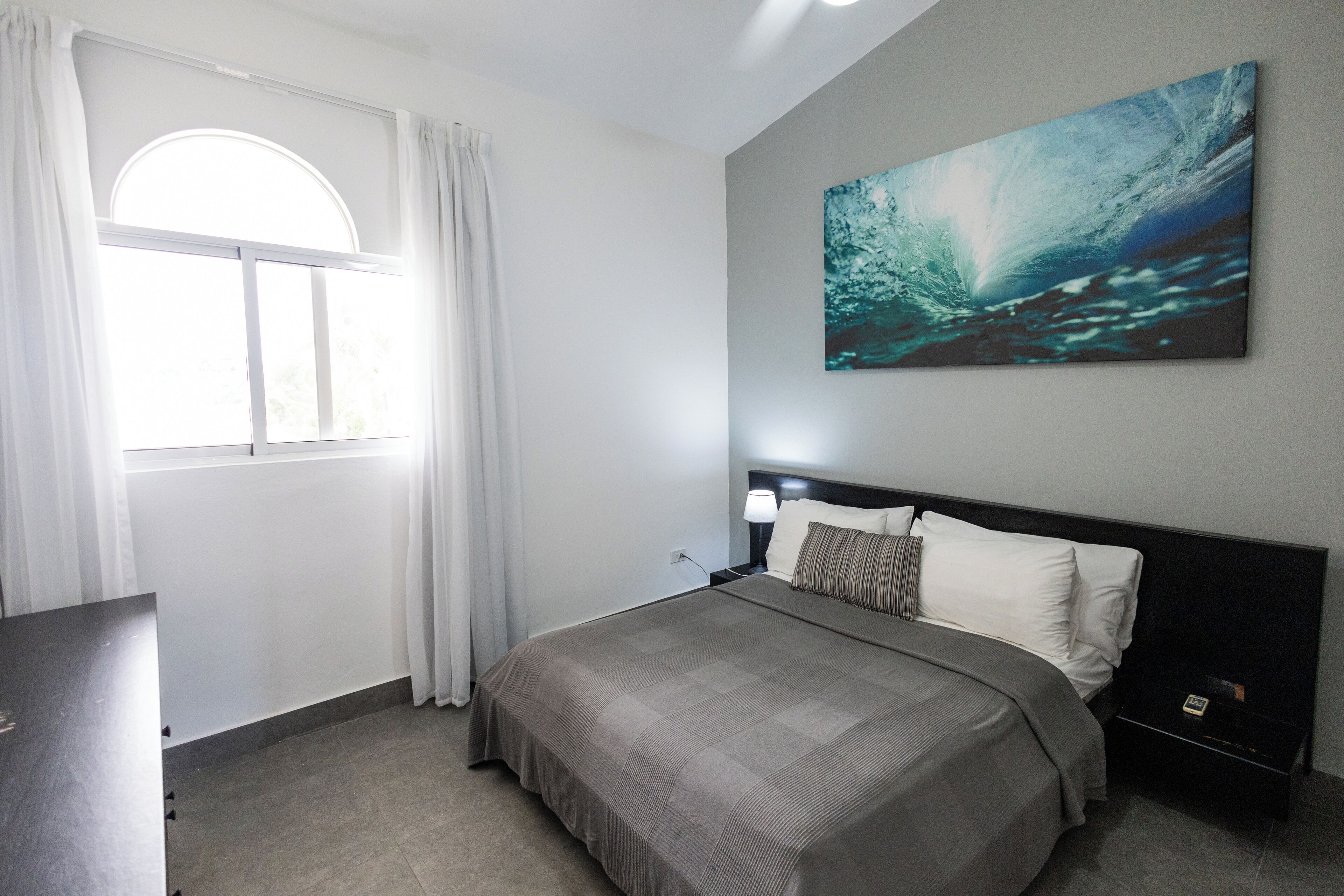 A modern bedroom with a large bed, a nightstand, a lamp, and a painting of a wave on the wall.