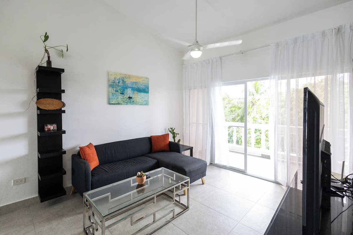 Una sala moderna con un sofá seccional, una mesa de centro de vidrio, un mueble de televisión negro, un ventilador de techo y un ventanal con cortinas ligeras.