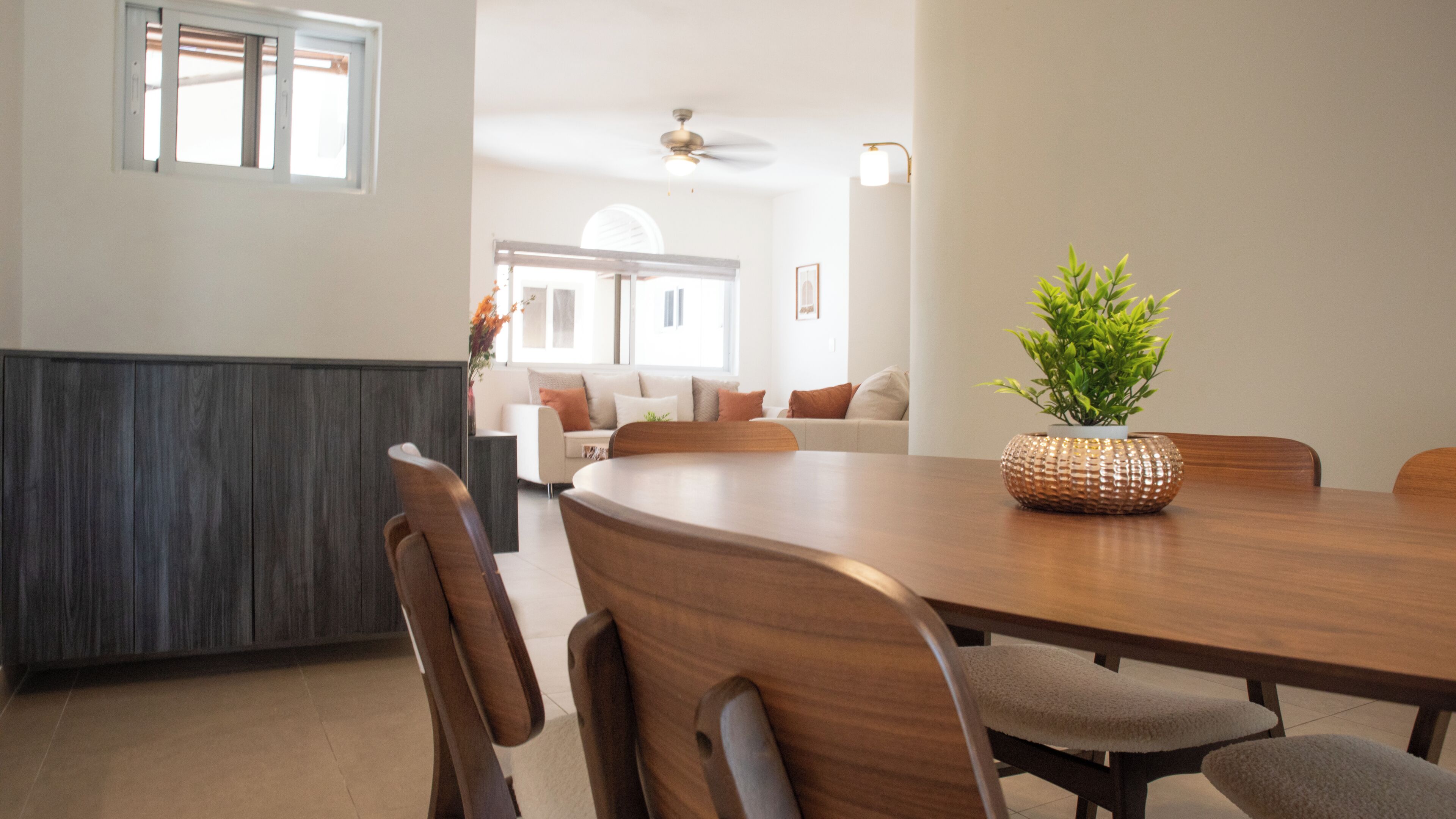 Een eetgedeelte met een houten tafel, stoelen, een plant en uitzicht op een woonkamer met een bank en een raam.