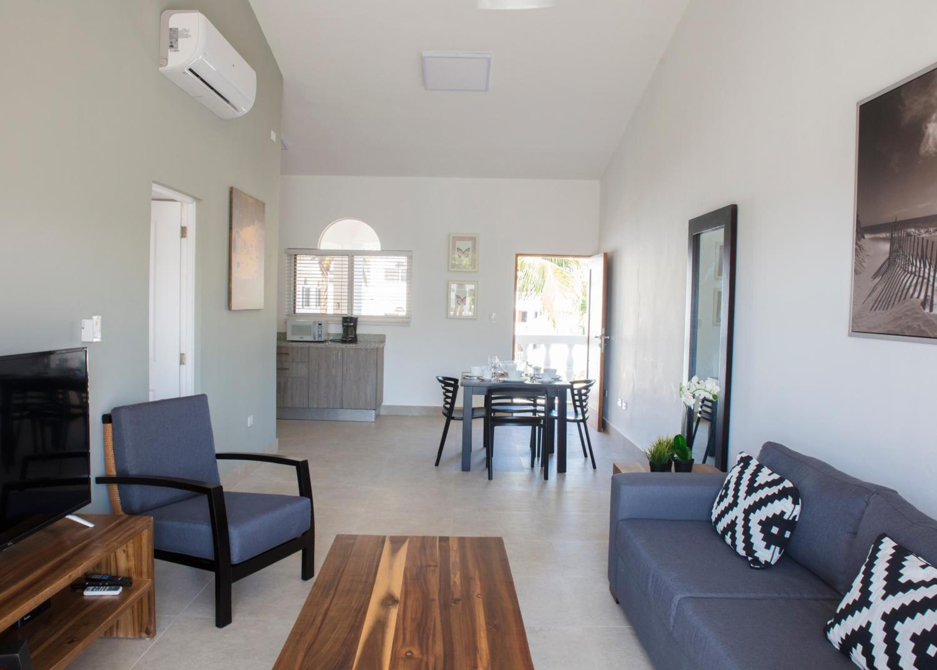 A modern living room with a TV, sofa, and coffee table. There is a kitchen area with a dining table and chairs.