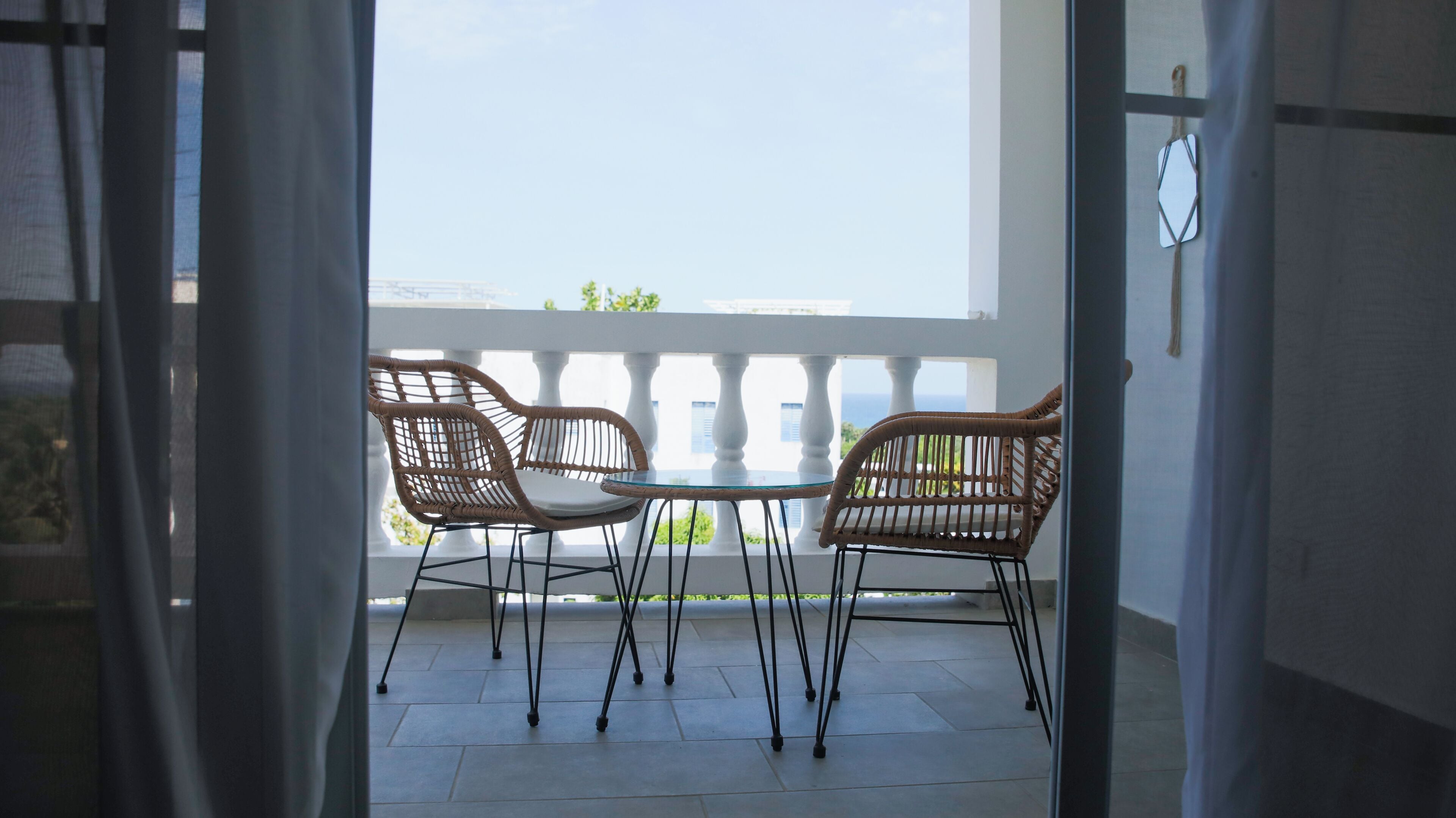 A balcony with two wicker chairs and a small round table, overlooking a white building and greenery.