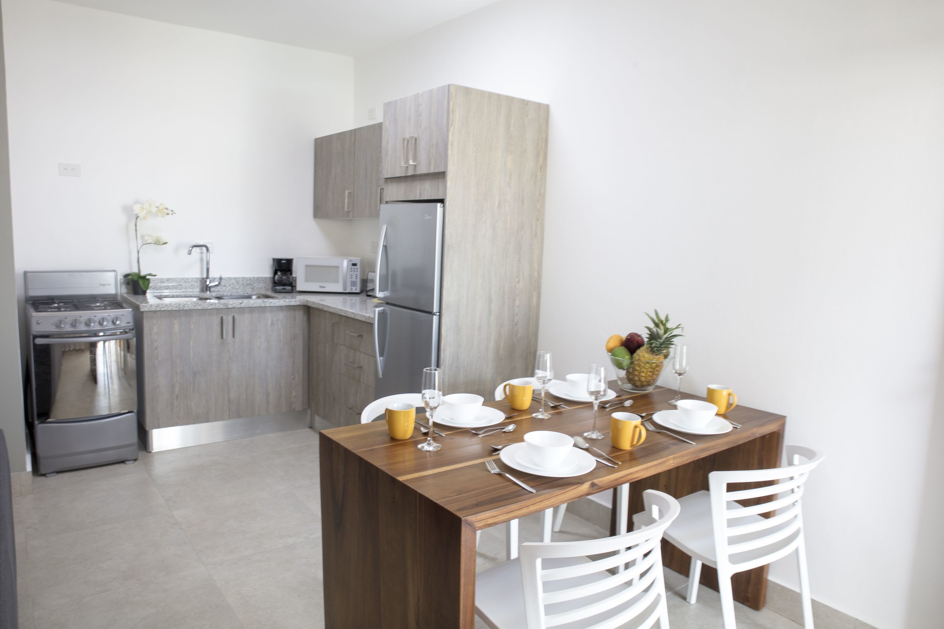 A modern kitchen with grey cabinets, a wooden dining table set with white plates, yellow cups, and a fruit bowl.