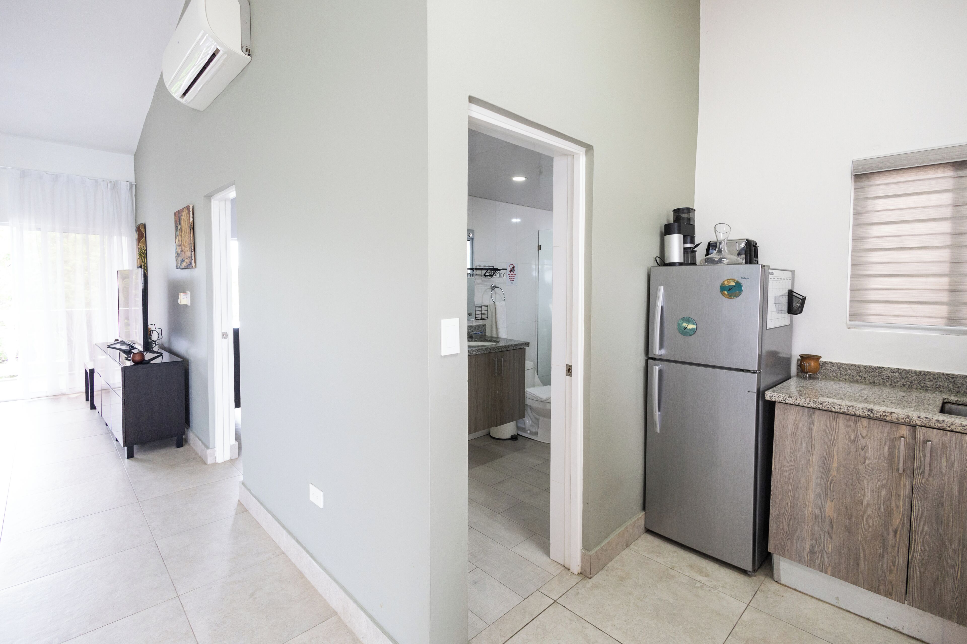 A modern kitchen with a refrigerator, a sink, and a countertop. There is a window with blinds and a door leading to another room.