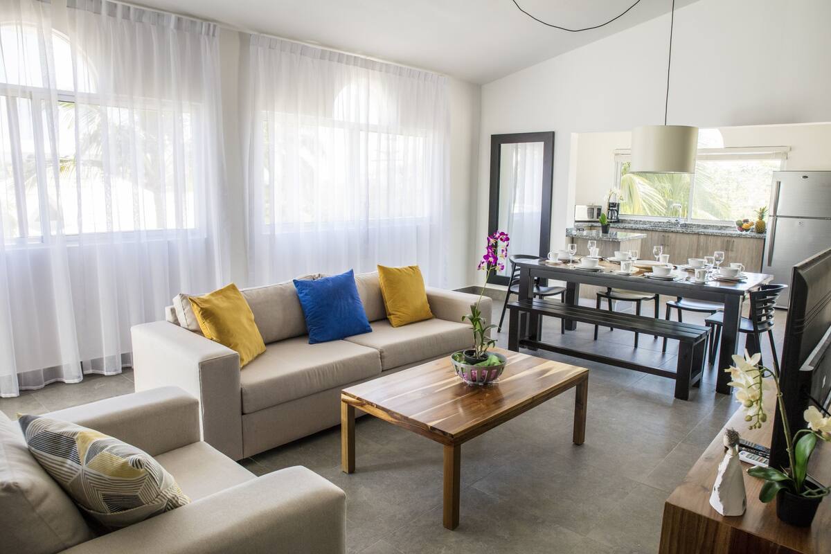 Una sala de estar moderna con sofá, mesa de centro y comedor, con una cocina al fondo.