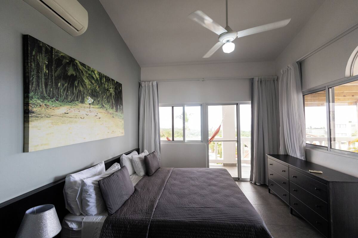 Un dormitorio con una cama grande, un ventilador de techo, un televisor y un balcón con cortinas.