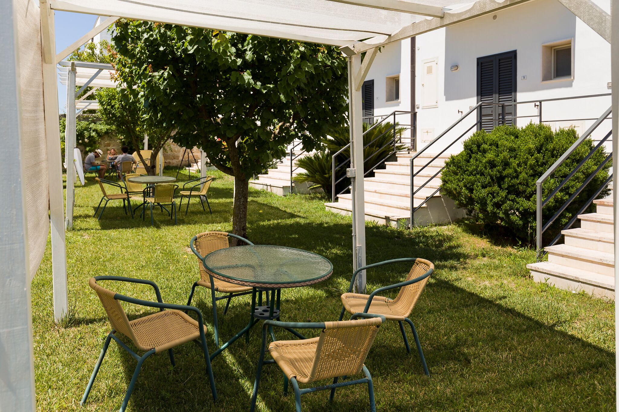 An outdoor patio with a round glass table and wicker chairs, surrounded by a white pergola.
