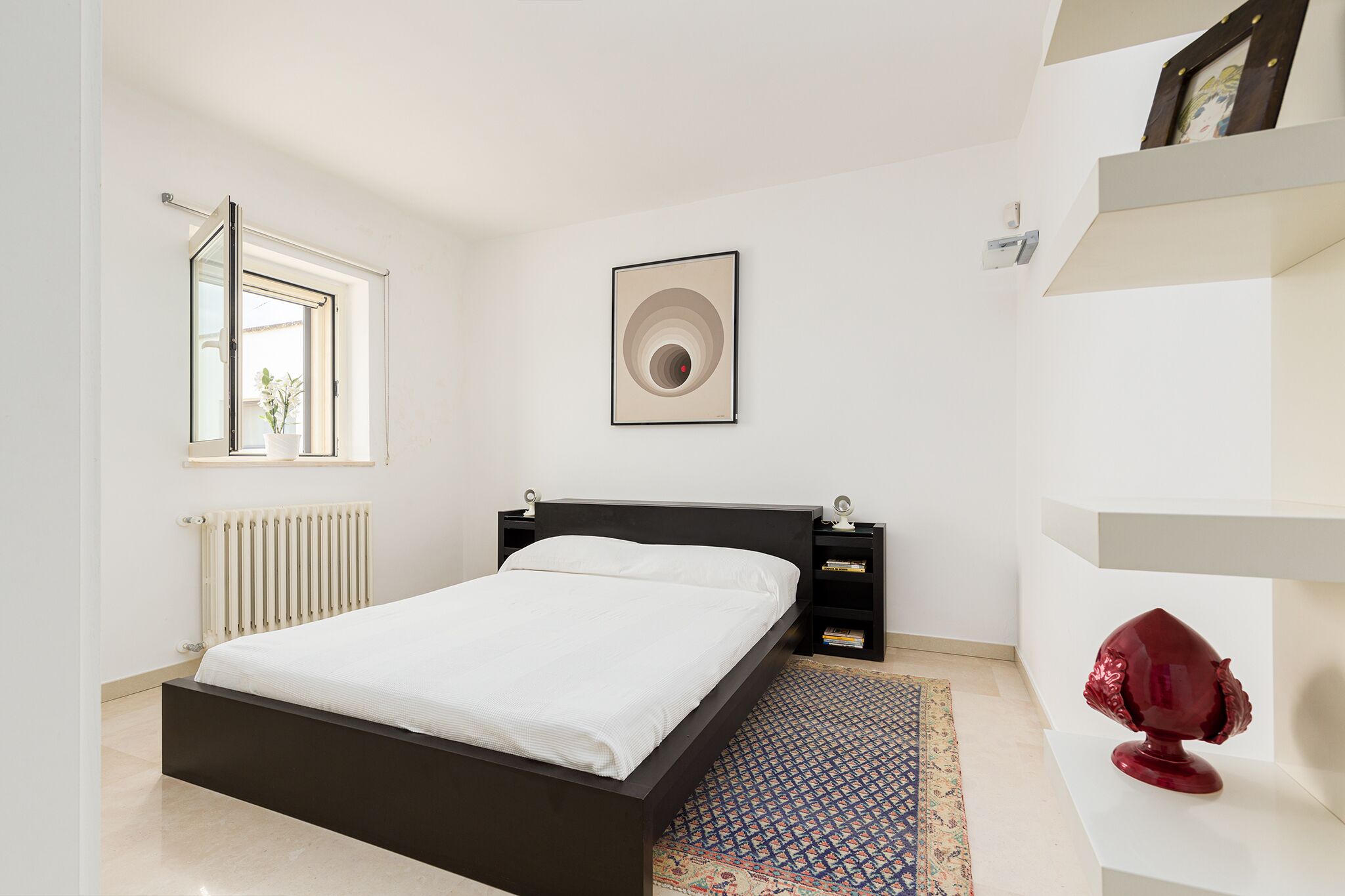 Un dormitorio moderno con una cama negra, un radiador blanco, una ventana y una escultura decorativa.