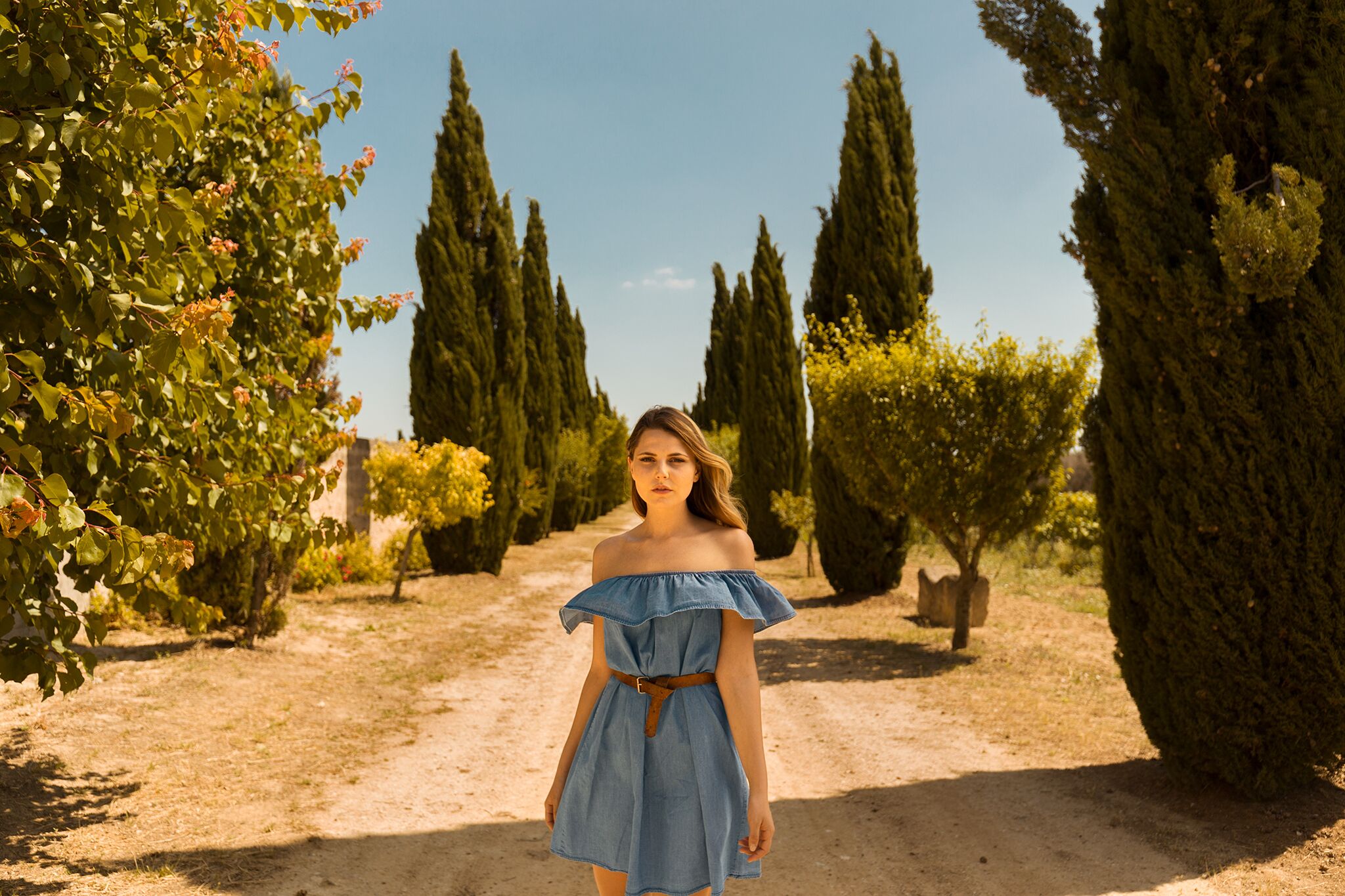 Eine Frau in einem blauen Kleid steht auf einem Feldweg, umgeben von Bäumen.