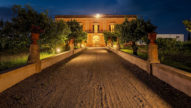 A building with a central entrance, flanked by trees and illuminated by street lamps. - Tenuta Giardini Nuovi (Novoli)