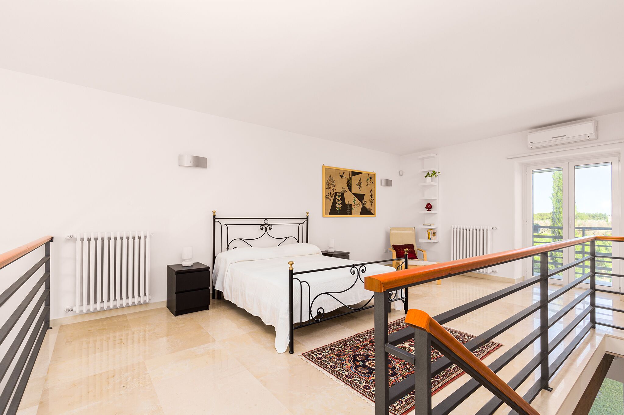 Quarto com cama, mesa de cabeceira, radiador, janela e corrimão de escada.