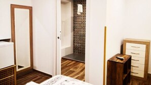 Une chambre avec un plancher en bois, un lit blanc, une commode en bois et une porte donnant sur une salle de bain dont le mur de la douche est décoré de motifs.