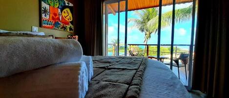 A hotel room with a large bed, a painting on the wall, and a view of palm trees through the window.