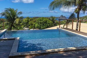 En swimmingpool med træterrasse, omgivet af palmer og udsigt til havet.