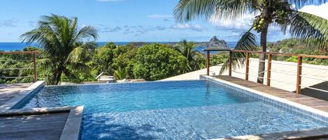 A swimming pool with a wooden deck, surrounded by palm trees and a view of the ocean.