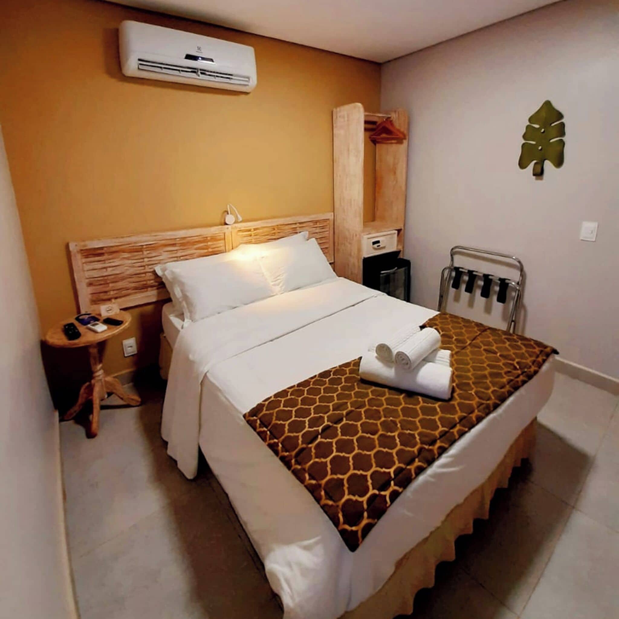 A hotel room with a bed, a bedside table, a small table with a lamp, an air conditioning unit, a towel rack, and a wall decoration.