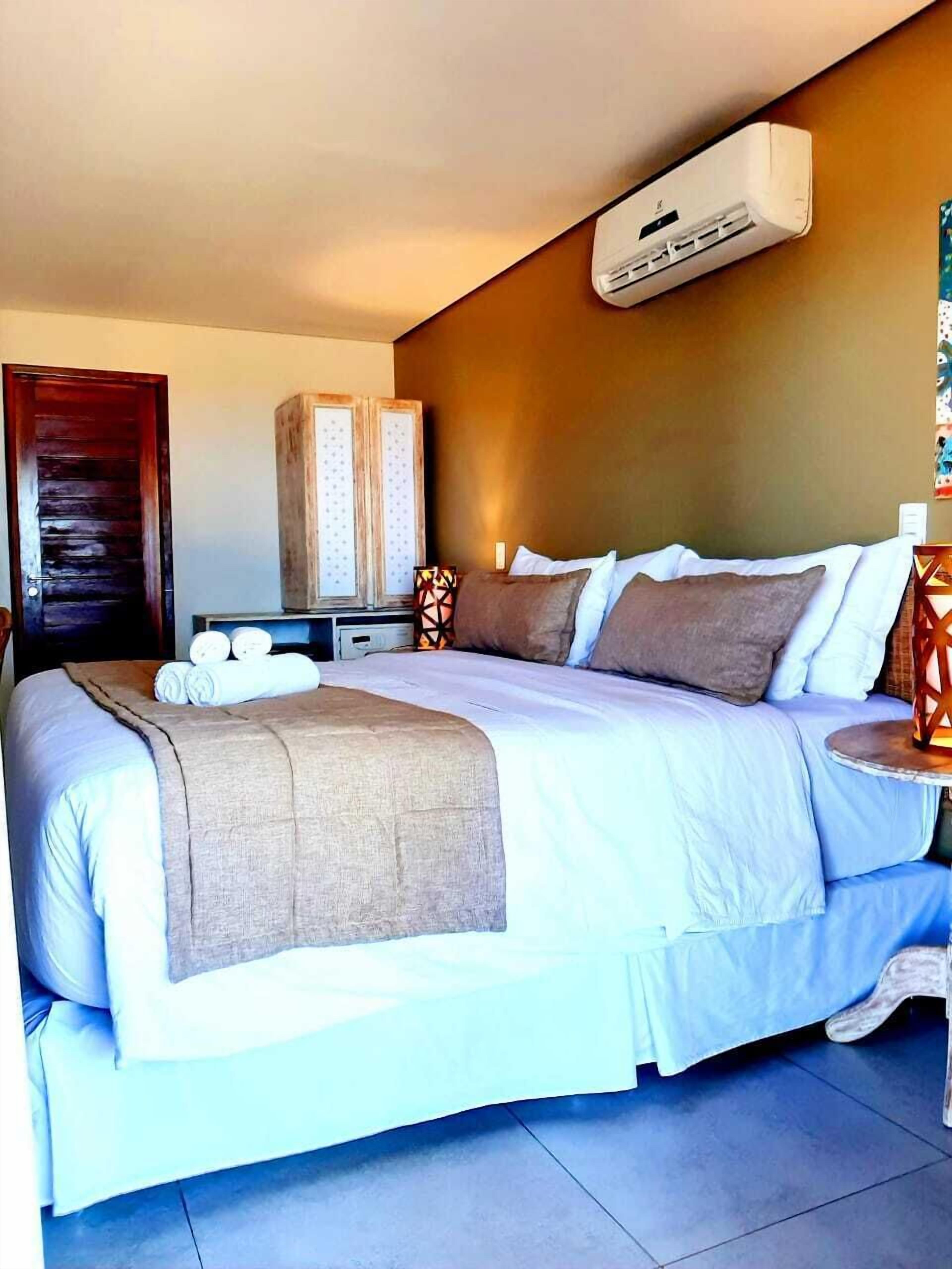 A hotel room with a large bed, a bedside table, an air conditioning unit, and a wall-mounted shelf with decorative items.