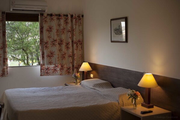 A hotel room with a bed, two bedside lamps, a nightstand, a framed picture on the wall, and a window with patterned curtains. - Hotel Portal de Gravatá (Gravata)