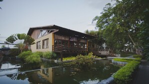 Un edificio de madera tradicional con un estanque grande en primer plano, rodeado de abundante vegetación.