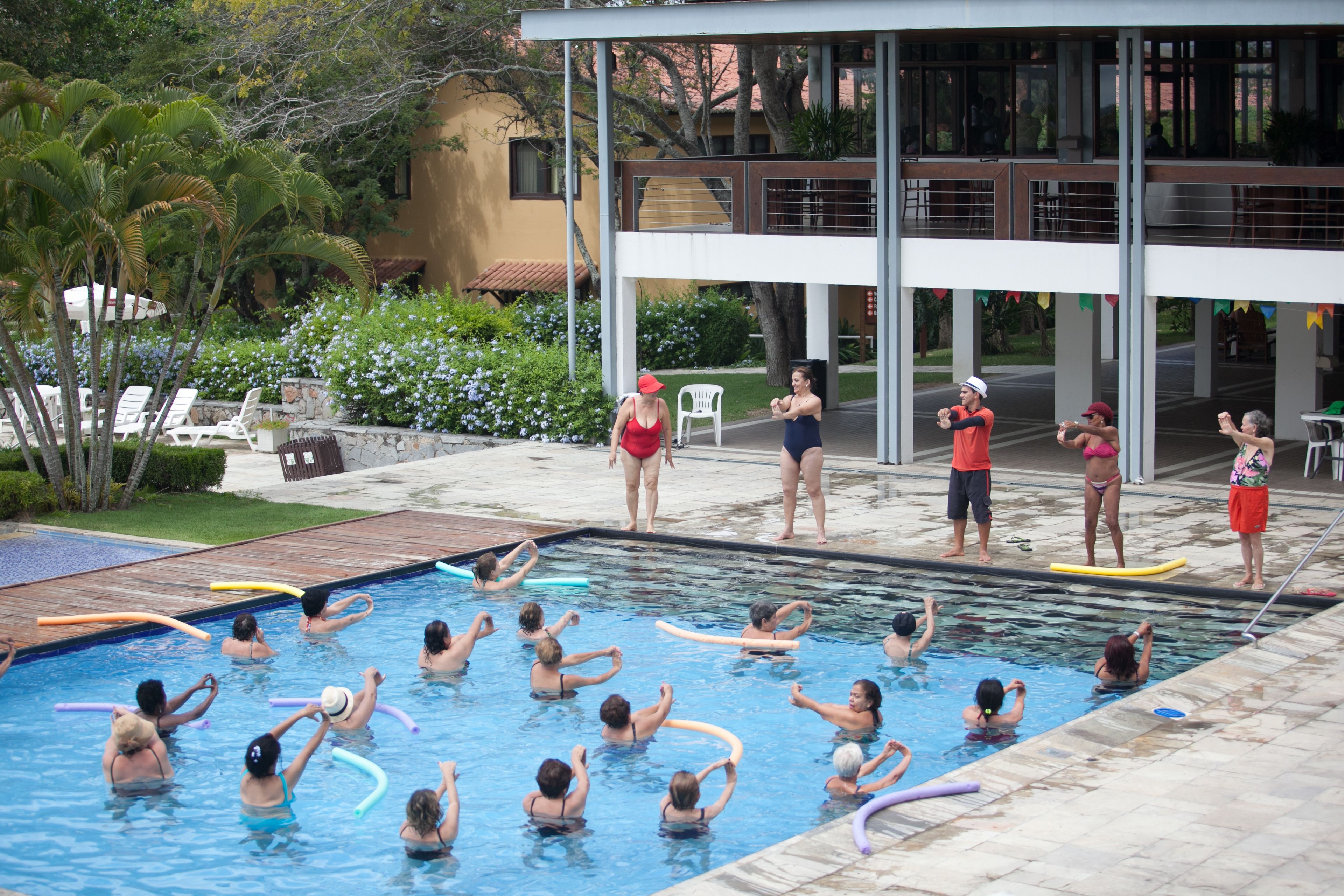 Uma área de piscina com várias pessoas praticando atividades aquáticas, cercada por um edifício com varanda e área de estar externa.