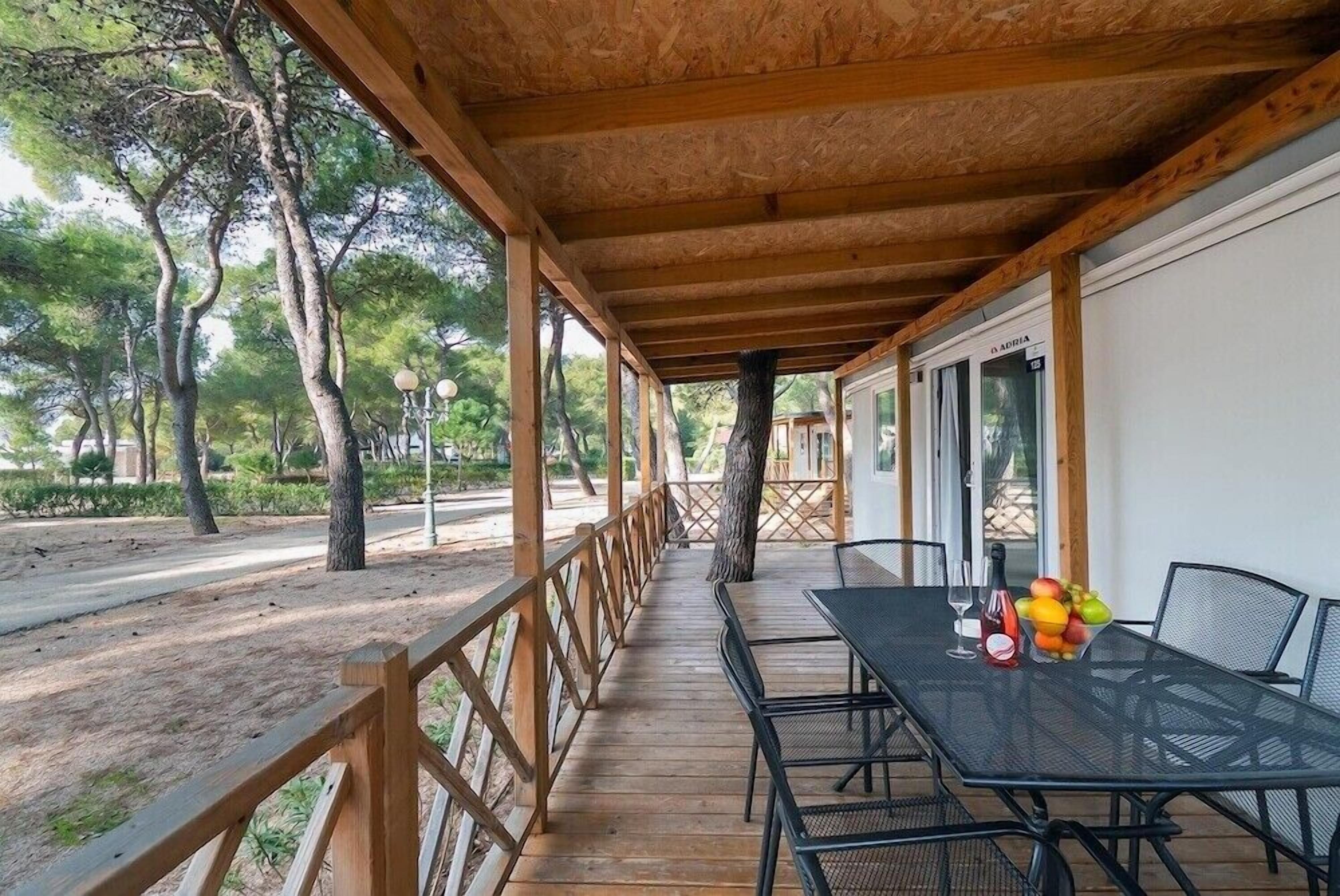 Een overdekt terras buiten met een tafel en stoelen, omgeven door bomen en een houten hekwerk.
