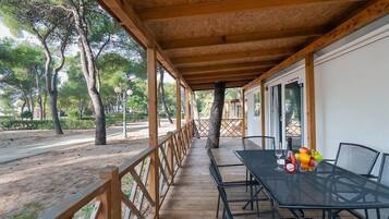 Een overdekt terras buiten met een tafel en stoelen, omgeven door bomen en een houten hekwerk.