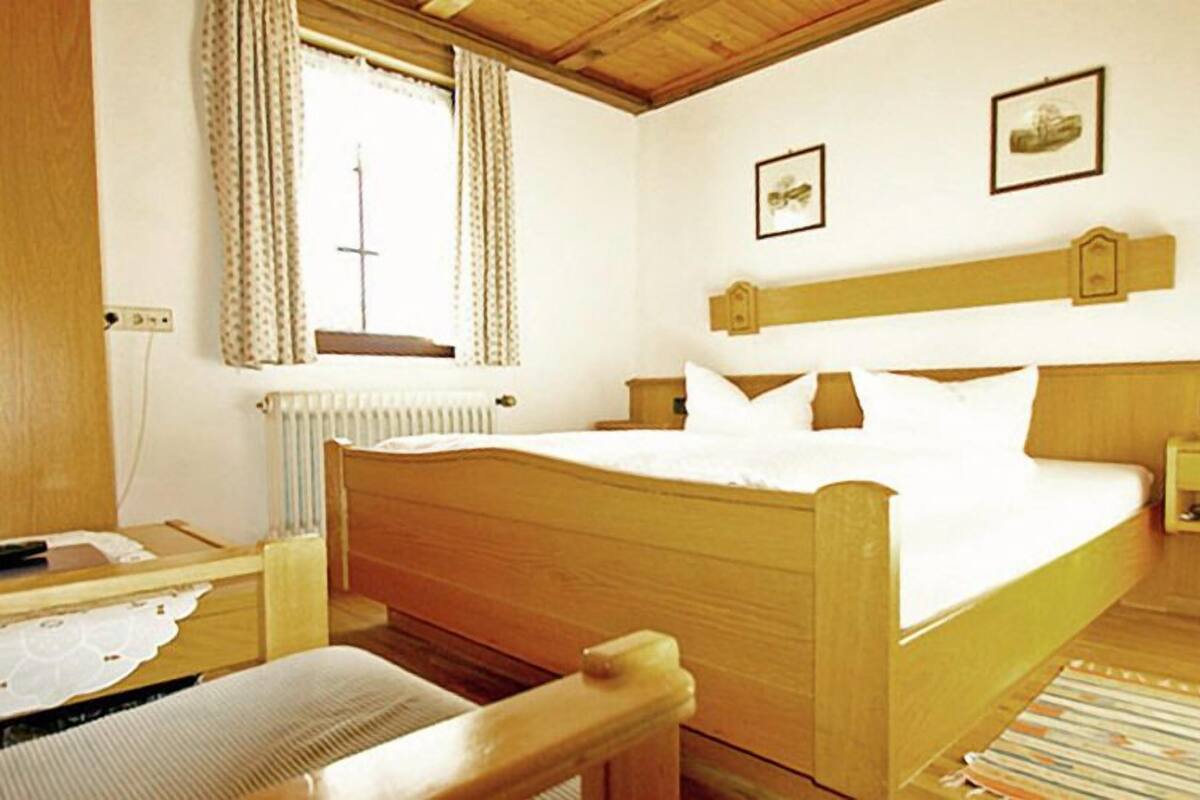 Ein hölzernes Bett mit weißer Bettwäsche, ein Nachttisch, ein Stuhl und ein Fenster mit Vorhängen.