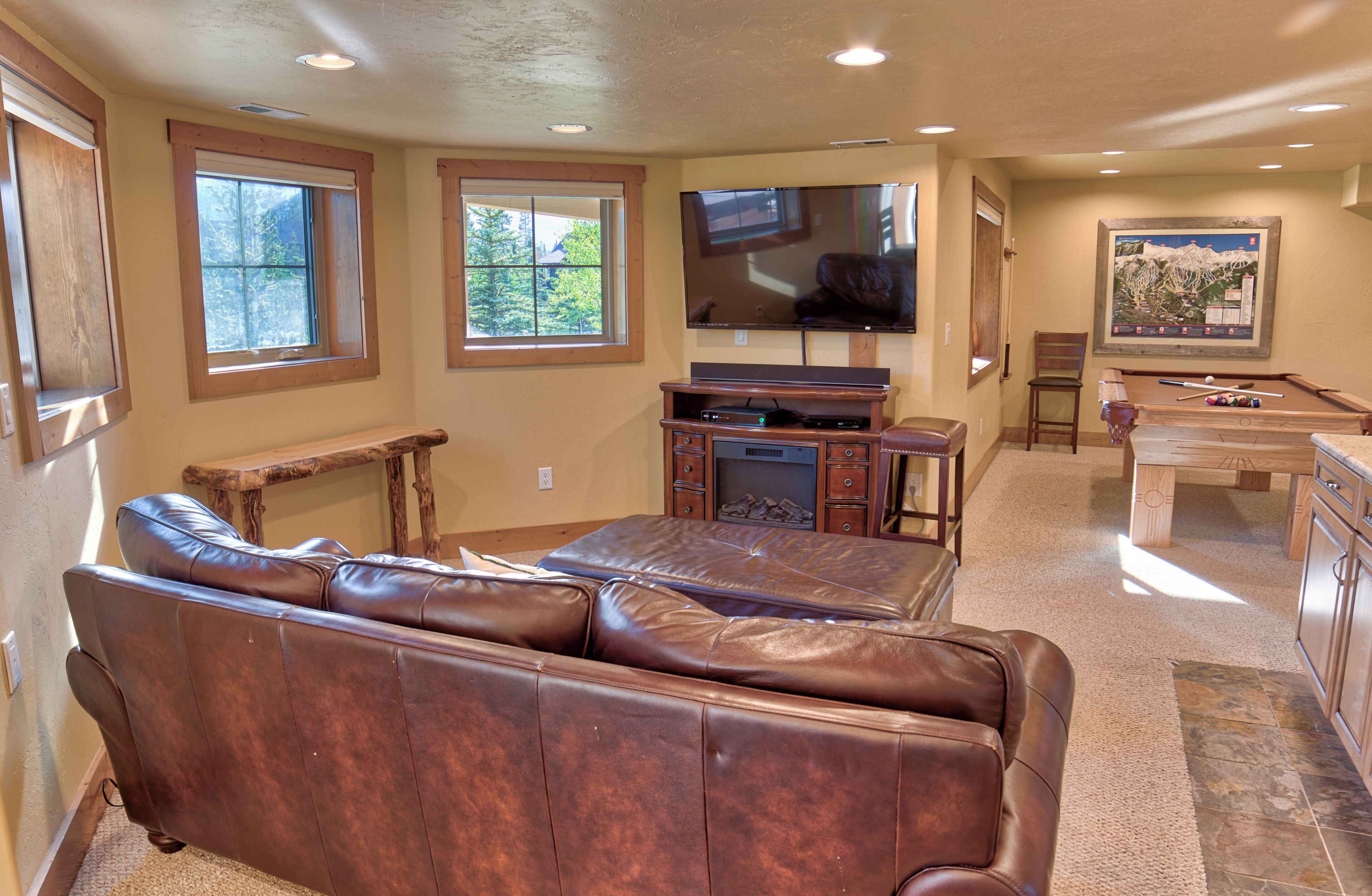 A spacious living room with a large leather sectional sofa, a flat-screen TV mounted above a wooden entertainment center, and a billiards table in the background.
