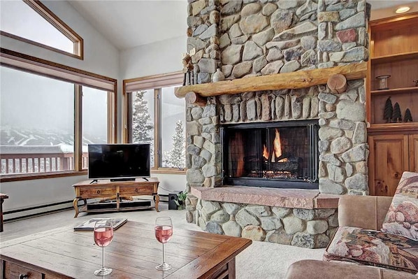 A cozy living room with a stone fireplace, a television on a wooden stand, and a table with two glasses of wine.