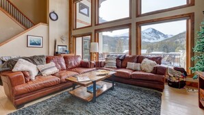 Un vaste salon doté d’un grand canapé d’angle en cuir marron, d’une table basse en bois et offrant une vue sur des montagnes enneigées grâce à de grandes fenêtres.