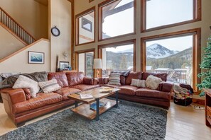 Ein geräumiges Wohnzimmer mit einem großen braunen Ledersofa, einem Holztisch und Blick auf schneebedeckte Berge durch große Fenster.