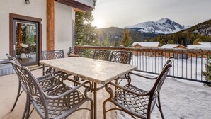 Un balcon avec une table et des chaises, une vue sur des montagnes enneigées et un bâtiment en bois doté d’une porte vitrée.