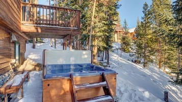 A wooden cabin with a hot tub, wooden deck, and snow-covered ground.