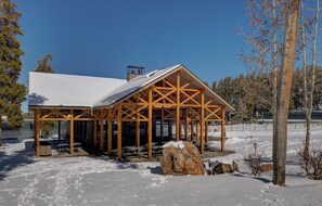 A wooden pavilion with a snow-covered roof, surrounded by snow-covered ground and trees.